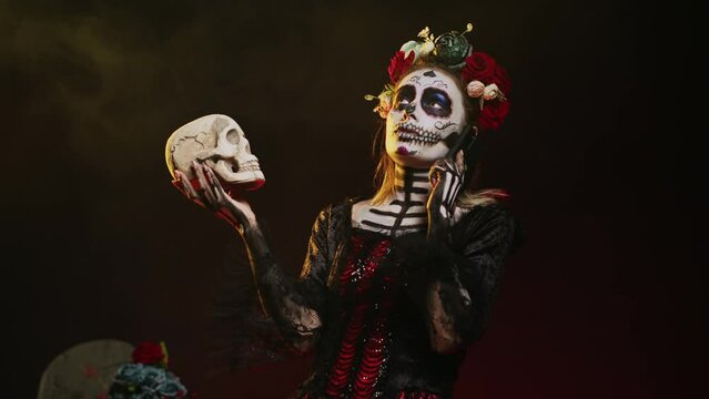 Mexican horror goddess on phone call holding skull, talking on smartphone line and acting creepy in studio. Answering telephone for remote chat, wearing holy mexican entity costume. Handheld shot.