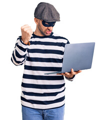 Young handsome man wearing burglar mask using laptop annoyed and frustrated shouting with anger, yelling crazy with anger and hand raised