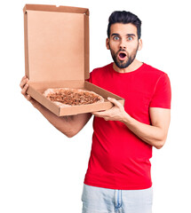 Young handsome man with beard holding delivery cardoboard with italian pizza scared and amazed with open mouth for surprise, disbelief face