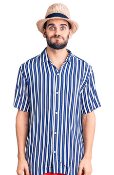 Young handsome man with beard wearing summer hat and striped shirt puffing cheeks with funny face. mouth inflated with air, crazy expression.