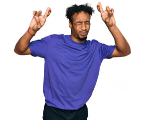 Fototapeta premium Young african american man with beard wearing casual purple t shirt gesturing finger crossed smiling with hope and eyes closed. luck and superstitious concept.