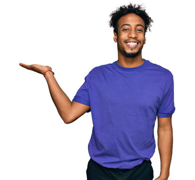 Young African American Man With Beard Wearing Casual Purple T Shirt Smiling Cheerful Presenting And Pointing With Palm Of Hand Looking At The Camera.