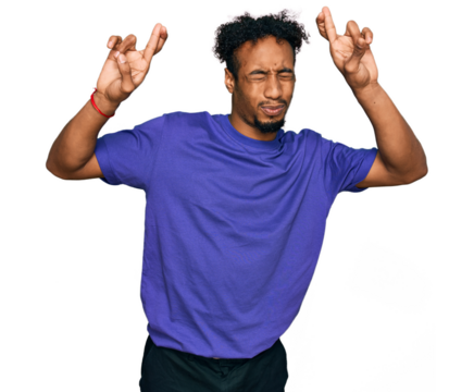Young african american man with beard wearing casual purple t shirt gesturing finger crossed smiling with hope and eyes closed. luck and superstitious concept.