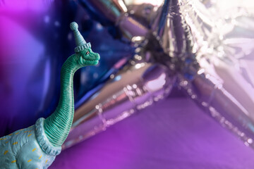 Offbeat party dinosaur wearing a party hat in front of shiny silver, purple and blue party balloons 