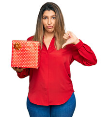 Beautiful brunette woman holding gift with angry face, negative sign showing dislike with thumbs down, rejection concept