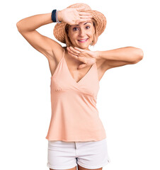 Young blonde woman wearing summer hat smiling cheerful playing peek a boo with hands showing face....