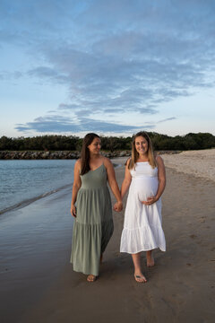 lesbian couple walking on the beach holding baby bump