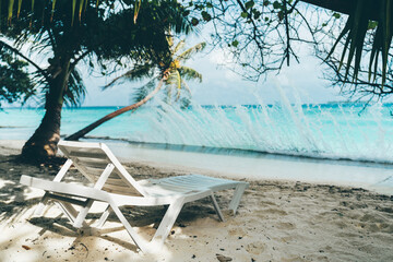A single white beach daybed in front of a waterfront with splashes of turquoise water; an empty...