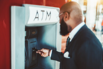 A side view of an elegant bearded bald black man entrepreneur in a suit and glasses with a selective focus on his hand inserting a credit card into an outdoor bank ATM to withdraw some cash
