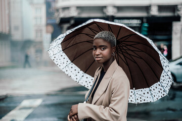 An outdoor portrait of an elegant youthful black female in a beige autumn overcoat and with short hair painted white, hiding from the rain under a big spotted brown umbrella on the street