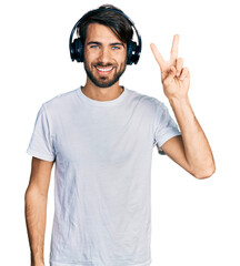 Hispanic man with blue eyes listening to music using headphones smiling with happy face winking at...