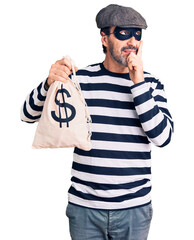 Middle age handsome man wearing burglar mask holding money bag serious face thinking about question...