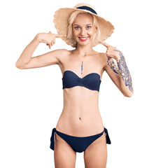 Young blonde woman with tattoo wearing bikini and summer hat smiling cheerful showing and pointing with fingers teeth and mouth. dental health concept.