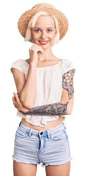 Young blonde woman with tattoo wearing summer hat looking confident at the camera with smile with crossed arms and hand raised on chin. thinking positive.
