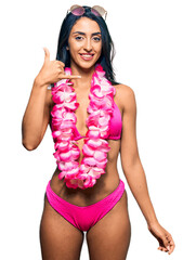 Beautiful hispanic woman wearing bikini and hawaiian lei smiling doing phone gesture with hand and...