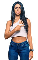 Beautiful hispanic woman wearing casual clothes pointing aside worried and nervous with forefinger,...
