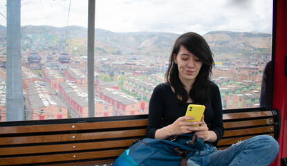 Beautiful Hispanic young woman sitting in the cable car cabin checking her mobile phone
