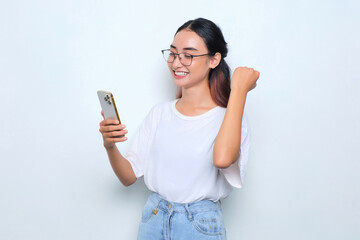 Excited young Asian girl in white t-shirt using smartphone and doing winner gesture isolated on...