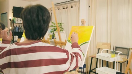 A talented Asian boy concentrates skill lessons on acrylic color picture painting on canvas in an art classroom and creatively learns with a female teacher at the elementary school studio education.