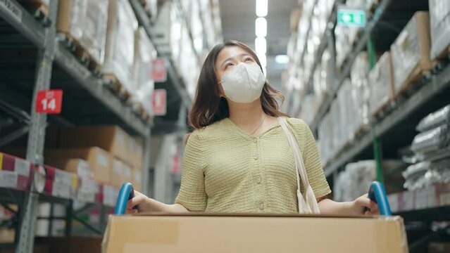 Asian woman wearing medical mask and walking in store steering shopping trolley and looking around at shelves with goods in furniture warehouse.