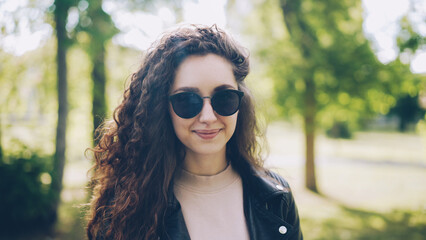 Portrait of attractive young woman in sunglasses and trendy leather jacket looking at camera and smiling standing in city park on summer day. People and sunlight concept.