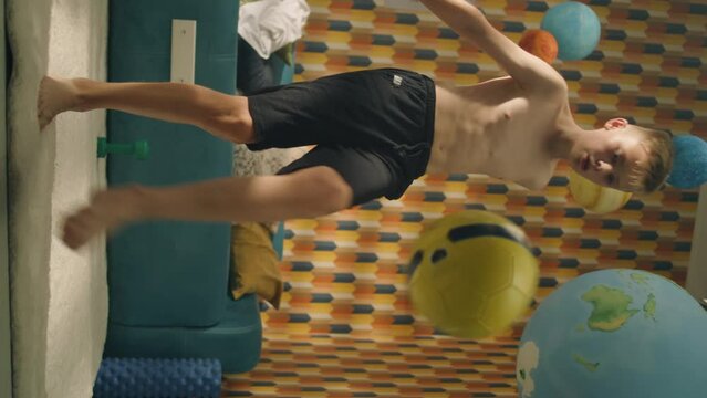 Vertical video shot of enthusiastic young football player trains controls a soccer ball in his room. Football fan practises football before the match. Teenage boy with bare upper body dribbles a ball.