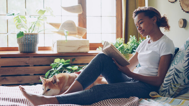 Pretty girl African American student is reading book and caressing pet dog expressing love and care sitting on bed in the house. Youth, hobby and domestic animals concept. - Powered by Adobe