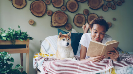 Smiling African American student lovely girl is reading book on bed at home while her pet dog is lying near her. Hobby, leisure, obedient animals and interior concept.