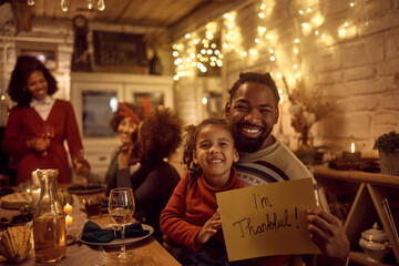 Happy black father and daughter holding placard with 'I'm thankful' while celebrating Thanksgiving with their extended family and looking at camera.