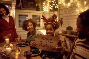 Happy black girl holding placard with Happy Thanksgiving message while having family dinner at dining table and looking at camera,