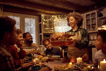 Mature black woman serving traditional Thanksgiving turkey during family meal at dining table.