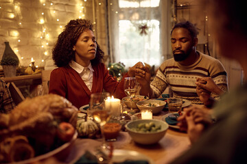 Black couple holding hands and praying while celebrating Thanksgiving with their family at dining table.