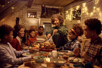 Happy African American grandmother carving Thanksgiving turkey during family lunch at dining table.