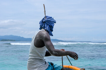A Melanesian man sitting on the bow of a fishing boat wearing head wrap scarf with a large feather...