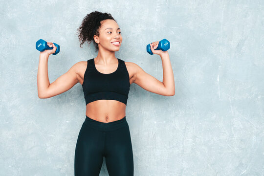 Fitness smiling black woman in sports clothing with curls hairstyle.She running and jogging . Young beautiful model with perfect tanned body. Female holding dumbbells in studio near grey wall