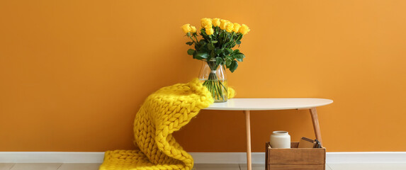 Vase with beautiful yellow roses and plaid on table near orange wall