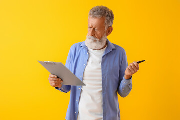 Thoughtful mature man with pen and clipboard on yellow background