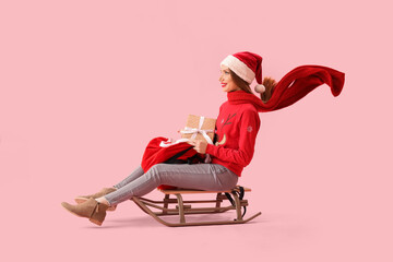 Beautiful young woman in Santa hat with bag of gifts and sledge on pink background - obrazy, fototapety, plakaty