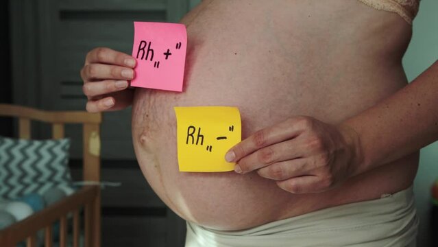 A pregnant woman holds multi-colored pieces of paper with the inscription positive and negative Rh factor against the background of her large belly. Rhesus conflict concept