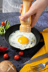 female hands using a wooden mill for spices prays peppers on fried eggs in a cast-iron skillet