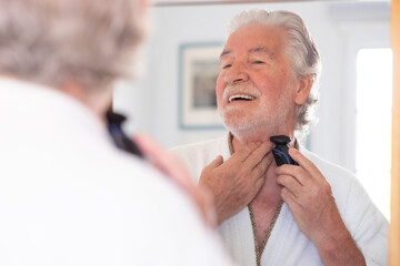 Close up portrait of a handsome senior man in bathrobe shaving beard with an electric razor looking...