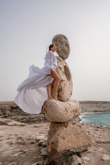 A woman stands on a stone sculpture made of large stones. She is dressed in a white long dress,...