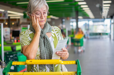 Senior woman in the supermarket checks her grocery receipt looking worried about rising costs - elderly lady pushing shopping cart, consumerism concept, rising prices, inflation - obrazy, fototapety, plakaty