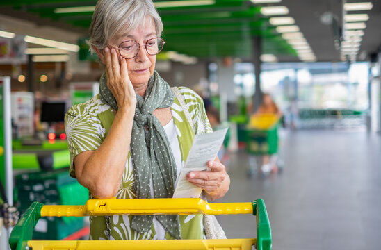 Senior woman in the supermarket checks her grocery receipt looking worried about rising costs - elderly lady pushing shopping cart, consumerism concept, rising prices, inflation
