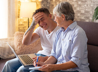 Young boy sitting on sofa at home helping senior grandmother surf with laptop on the web while she takes notes. Younger generations care for older relatives are teaching computers to use