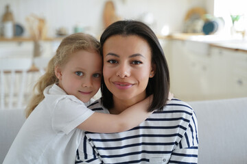Little adopted daughter hugging foster mother, sitting on sofa at home. Happy motherhood, adoption