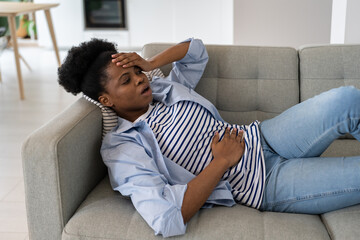 Sick young African American woman having health problems holding head and belly during menstruation. Black girl lying in living room on couch suffering from pain associated with gynecological causes - obrazy, fototapety, plakaty