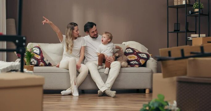 Happy young blonde woman mother wife with husband father sitting in new apartment with preschooler son having fun moving in process delighted parents carton boxes full of home stuff potted plats .