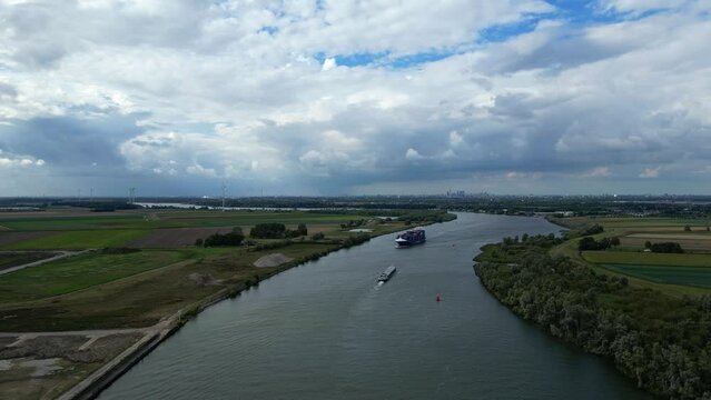 Steady and stable aerial shot of BG Onyx, a freight line container ship sailing across river with dramatic sky, vast landscapes and town of Zwijndrecht at the background