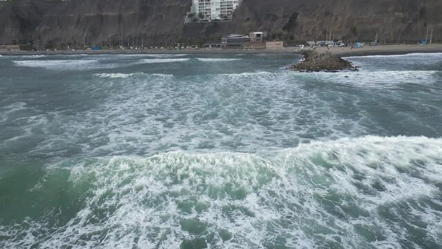 Slow motion aerial dolly shot of the sea with waves in front of the beach of miraflores lima in peru with rocks and building in the background